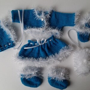 May include: A blue knitted baby outfit with white fur trim. The outfit includes a hat, cardigan, skirt, and booties.