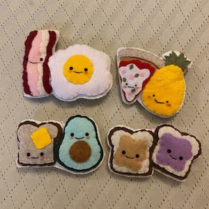 May include: A set of felt food patches featuring a fried egg, bacon, pizza slice, pineapple, avocado toast, and peanut butter and jelly toast. Each patch has a cute cartoon face.