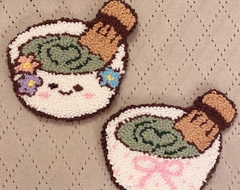 May include: Two handmade, tufted coasters. One coaster features a white bowl with a smiling face, flowers, and green matcha. The other has a pink bow. Both have a brown rim and a brown whisk.