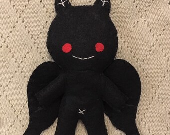 May include: Black felt plush toy with red eyes and wings. The toy is shaped like a small creature with a smiling face.