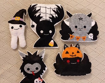 May include: Five felt Halloween character ornaments. A black and white deer with a spider web, a grey goat with a star and a banner that says "BOO", a black and white monster with a red tongue, a black and white spider with an orange pumpkin head, and a white ghost with a black hat.