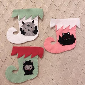 May include: Three felt Christmas stockings with different characters. The first stocking is white with green trim and a gray goat with black wings. The second stocking is pink with white trim and a black bat with white eyes. The third stocking is green with red trim and a black werewolf with a red tongue.