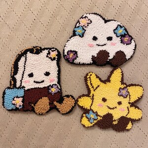 May include: Three punch needle coasters featuring a white cloud, a white mug, and a yellow star. Each character has a smiling face with pink cheeks, dark brown accents, and small colourful floral details in blue, pink, and purple yarn textures.