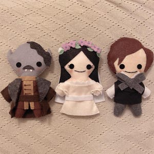 Puede incluir: Tres muñecas de fieltro, un monstruo gris, una muñeca de novia blanca con una corona de flores y una muñeca de pelo castaño que lleva un chaleco gris y pantalones negros.