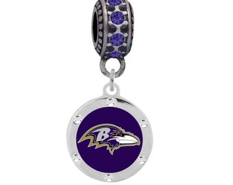 Może przedstawiać: Srebrny charm z logo Baltimore Ravens w kolorze fioletowym i złotym na okrągłym charmie z przezroczystymi cyrkoniami wokół krawędzi. Charm jest przymocowany do srebrnej koraliki z fioletowymi cyrkoniami.
