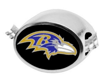 May include: A silver charm featuring a black background and a purple and gold Baltimore Ravens logo. The logo depicts a raven with a "B" on its chest.