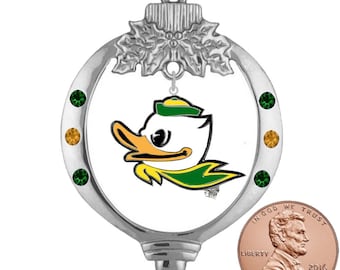 May include: Silver Christmas bauble featuring a green and yellow duck mascot with a green hat. The bauble has a silver ring with green and gold rhinestones. The bauble is hanging from a silver string.