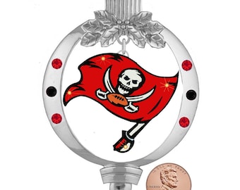 Puede incluir: Adorno navideño plateado con el logotipo de los Tampa Bay Buccaneers. El adorno es redondo con un logotipo rojo y blanco de una calavera de pirata y huesos cruzados con una pelota de fútbol. El adorno está decorado con diamantes de imitación rojos y negros.