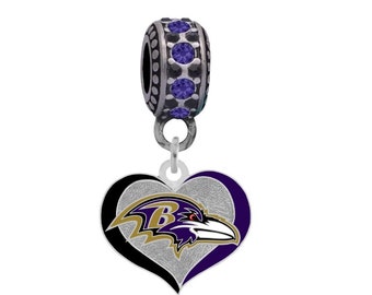 Puede incluir: Un charm plateado con un corazón morado, negro y dorado con el logotipo de los Baltimore Ravens. El charm tiene una cuenta plateada con diamantes de imitación morados.