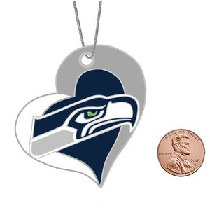 May include: A white and gray heart-shaped ornament with a blue and green Seattle Seahawks logo. The logo features a stylized eagle head with a green eye.