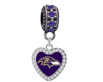 Puede incluir: Un charm plateado con un colgante en forma de corazón morado que presenta el logotipo de los Baltimore Ravens. El corazón está rodeado de diamantes de imitación transparentes y el charm tiene una cuenta de cristal morado.