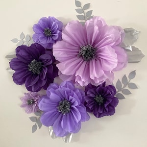 Pode incluir: Uma decora&ccedil;&atilde;o de parede com um grupo de flores de papel em tons de roxo e lavanda. As flores est&atilde;o dispostas em forma circular, com a flor maior no centro. As flores s&atilde;o feitas de papel de seda e t&ecirc;m uma textura delicada e ondulada.