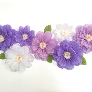 Puede incluir: Una colecci&oacute;n de siete grandes flores de papel en tonos de morado, lavanda y blanco. Las flores est&aacute;n dispuestas en una fila sobre un fondo blanco.