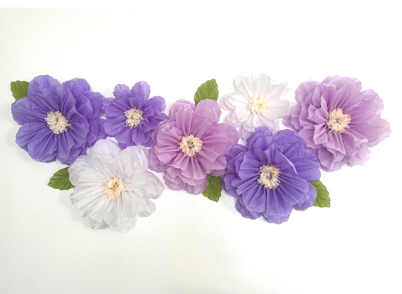 Puede incluir: Una colecci&oacute;n de siete grandes flores de papel en tonos de morado, lavanda y blanco. Las flores est&aacute;n dispuestas en una fila sobre un fondo blanco.