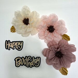 Puede incluir: Pancarta de Feliz Cumplea&ntilde;os con letras doradas brillantes y tres flores de papel de seda en tonos rosa y crema.
