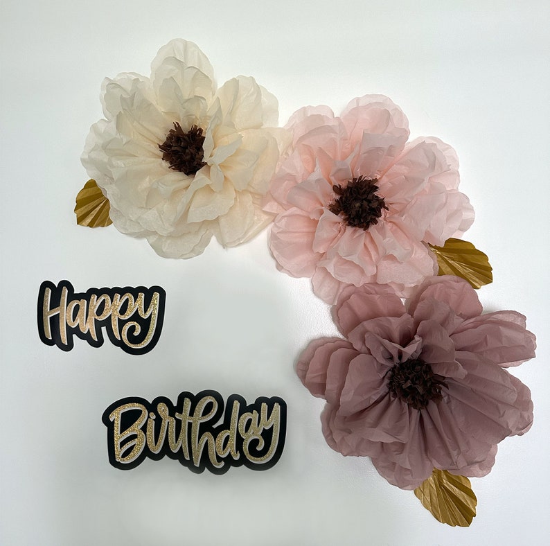 Puede incluir: Pancarta de Feliz Cumplea&ntilde;os con letras doradas brillantes y tres flores de papel de seda en tonos rosa y crema.