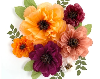 May include: A collection of layered paper flowers in shades of orange, peach, deep magenta, and white, featuring dark brown centers. The arrangement includes several green paper leaves and small vine-like sprigs with tiny leaves extending outward.