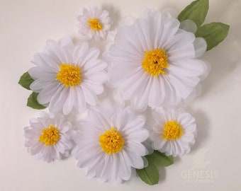 May include: A set of six white paper daisies with yellow centers. The daisies are arranged in a cluster on a white background. The largest daisy is in the center of the image, and the other daisies are arranged around it. There are also two green leaves in the image.