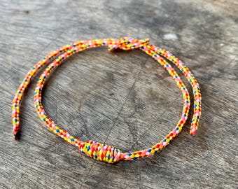 May include: A colorful braided rope bracelet with a knotted closure. The bracelet is made with yellow, red, pink, blue, and white threads.