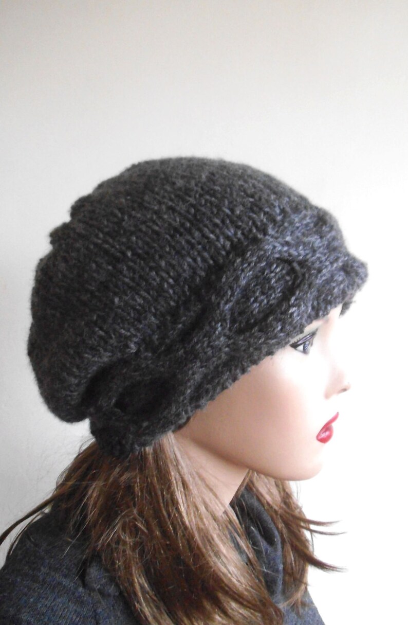 May include: A dark grey knitted beanie with a cable knit design. The beanie is slouchy and has a wide brim.