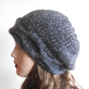 May include: A grey knitted beret with a cable knit design. The beret is worn on a mannequin with long brown hair.