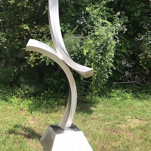 May include: A modern, abstract sculpture made of polished stainless steel. The sculpture features three curved pieces that intersect and create a dynamic, flowing form. The sculpture is mounted on a square base.