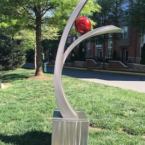 May include: A modern outdoor sculpture made of stainless steel with a red sphere at the top. The sculpture is on a grassy lawn in front of a building.