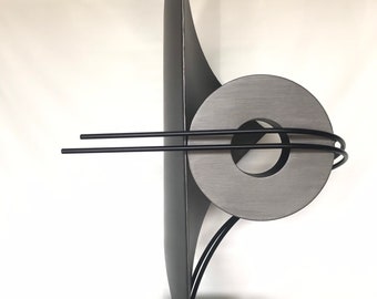 May include: Abstract metal sculpture with a circular form and a tall, thin, curved form. The sculpture is painted in a dark gray color and has a black base.