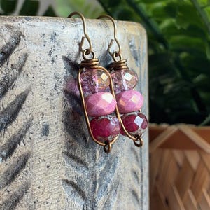 May include: A pair of dangle earrings featuring bronze-toned wire frames that encase three stacked, faceted beads in varying shades of pink and magenta. The earrings are suspended from simple bronze-toned metal hooks for pierced ears.