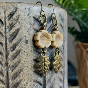 May include: Cream-coloured floral earrings with brass-coloured leaf charms. The earrings have a vintage aesthetic, with a flower-shaped bead and a brass leaf charm. The earrings are hanging in front of a textured, light brown background.