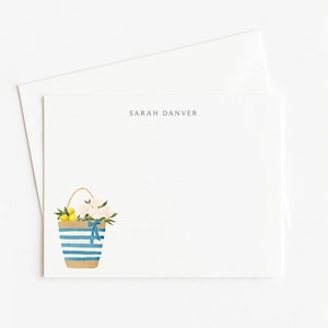 May include: White stationery with a watercolor illustration of a striped tote bag filled with lemons and flowers. The bag has a blue ribbon tied to the handle. The name "SARAH DANVER" is printed at the top of the stationery.