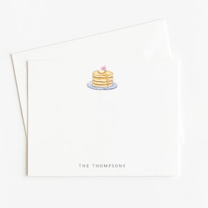 May include: White stationery card with a watercolor illustration of pancakes on a blue plate, topped with butter and a flower. The text "THE THOMPSONS" is printed in black at the bottom. Includes a matching envelope.