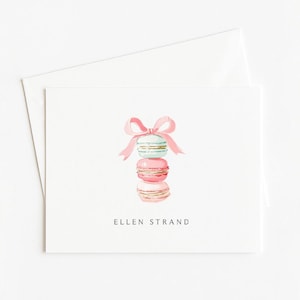 May include: A white greeting card with a watercolor illustration of three stacked macarons in pink and teal, tied with a pink bow. The card has the text "ELLEN STRAND" printed in black.