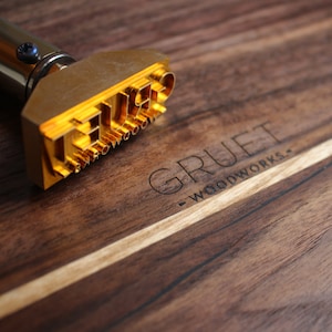 Puede incluir: Un hierro de marcar de metal dorado con el texto "THE" y "GRUE" en él. El hierro de marcar está sobre una superficie de madera con el texto "GRUET WOODWORKS" grabado en ella.