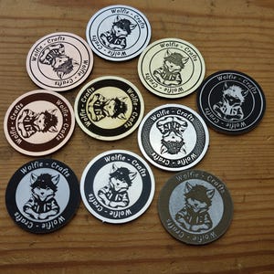May include: Ten round, silver, gold, and black metal tokens with a black and white image of a cartoon raccoon wearing a shirt and the text "Wolfie - Crafts" around the edge.