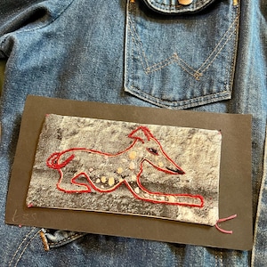 May include: A denim jacket with a black card featuring a red embroidered greyhound design. The artwork is stitched with red thread, and the background is a mottled grey. The jacket has a pocket and a button.