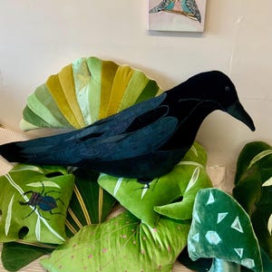 May include: A collection of decorative pillows in various shapes and colors. A large black crow pillow rests atop several green leaf-shaped pillows, a fan-shaped pillow, and a mushroom-shaped pillow. A small painting of two birds hangs in the background.