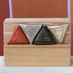 May include: Four triangular leather coin purses in different colors: orange, silver, green, and gold. The purses are arranged in a row on a wooden shelf.