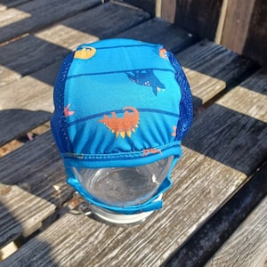 Puede incluir: Un gorro de natación azul con un estampado de dinosaurios. El gorro tiene un panel lateral de malla y está colocado sobre un recipiente de vidrio transparente. Los dinosaurios son de color naranja y el fondo es azul. El gorro está sobre una superficie de madera.