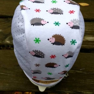 Peut inclure: Housse en tissu blanc ornée d'un motif de hérissons et de flocons de neige de dessin animé. La housse est enveloppée autour d'une structure en maille blanche. Les hérissons sont dans des tons de marron, gris et rose, avec des flocons verts et rouges.
