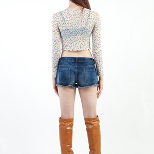 May include: A woman wearing a white floral crop top with long sleeves, blue denim shorts, and brown knee-high boots.