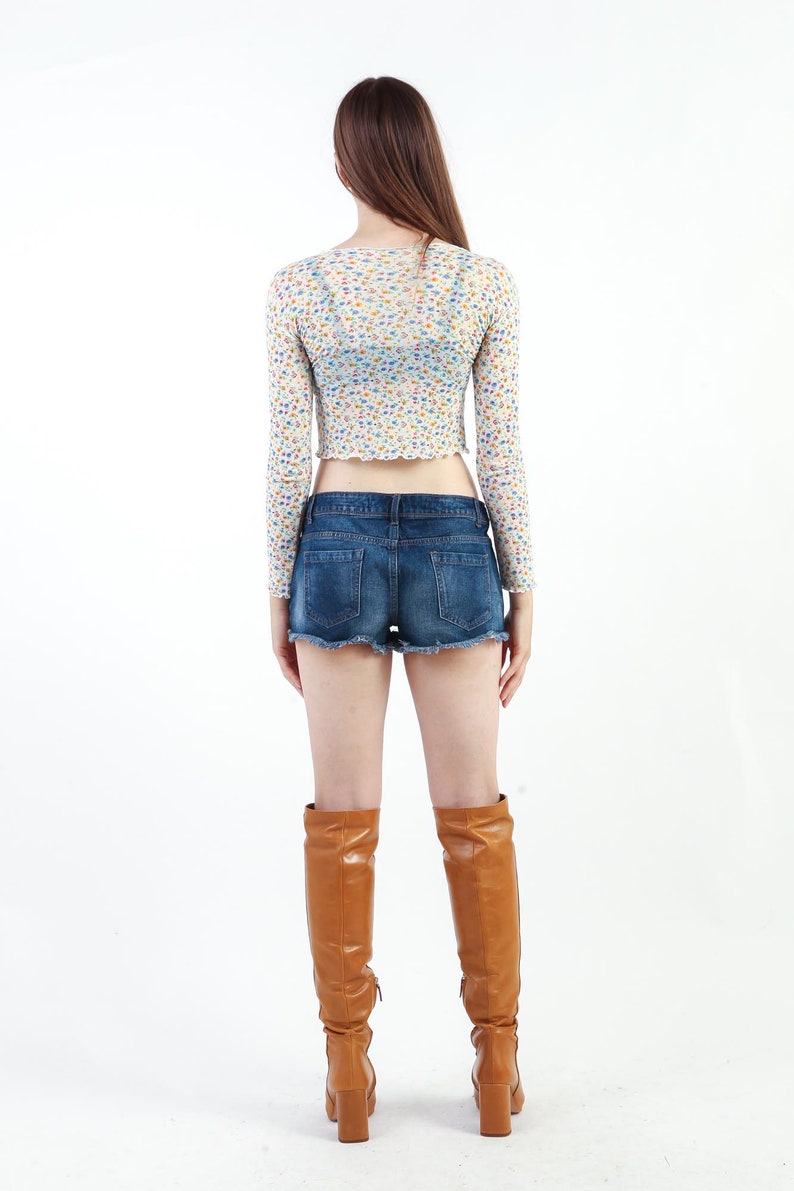 May include: A woman wearing a white floral crop top with long sleeves, blue denim shorts, and brown knee-high boots.