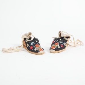May include: A pair of black espadrille shoes with colorful embroidered flowers and beads. The shoes have white laces that tie up the ankles.