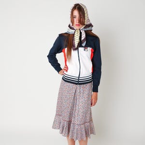 May include: A navy blue, white, and red zip-up track jacket with the Fila logo on the chest, paired with a floral print midi skirt featuring a ruffled hem and a headscarf with a multi-colored striped pattern tied around the head.