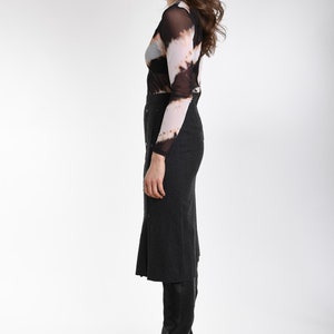 May include: A long-sleeved mesh top featuring a black, white, and tan tie-dye pattern paired with a dark gray, high-waisted midi skirt. The skirt includes a vertical row of buttons down the front and is worn with black leather knee-high boots.
