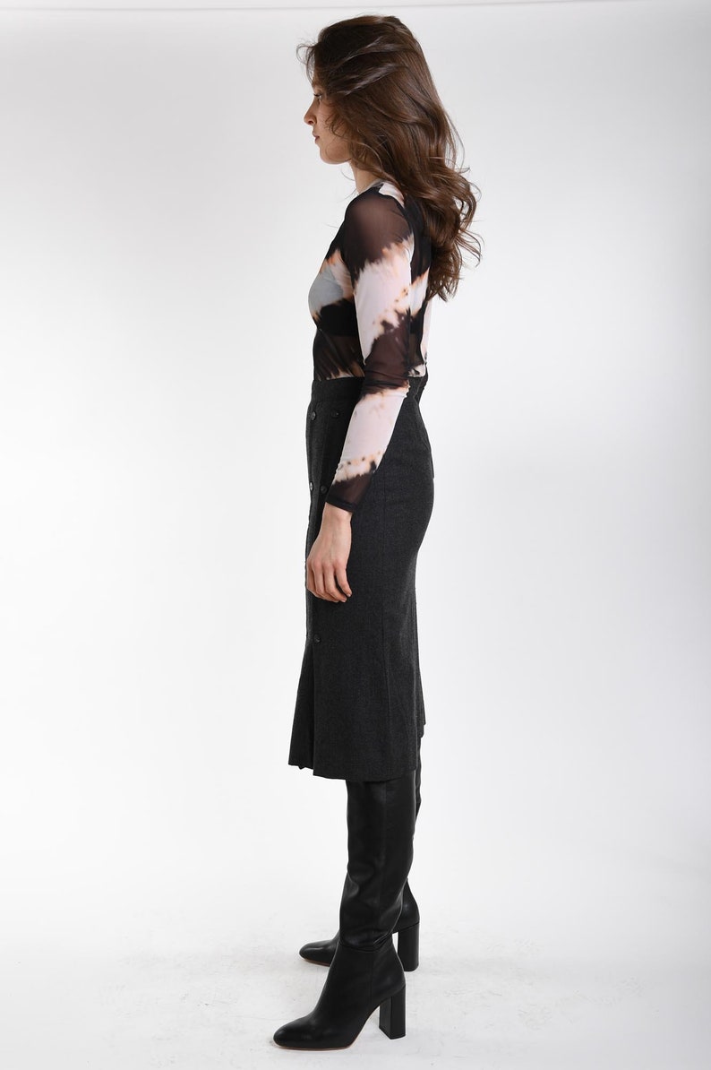 May include: A long-sleeved mesh top featuring a black, white, and tan tie-dye pattern paired with a dark gray, high-waisted midi skirt. The skirt includes a vertical row of buttons down the front and is worn with black leather knee-high boots.