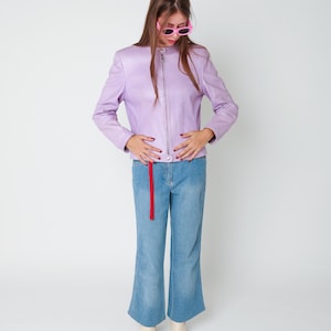 May include: A lavender leather jacket with a silver front zipper and a mandarin collar, paired with light blue denim jeans featuring a red fabric strip hanging from the waist and subtle embroidery at the hem, plus cream-colored ankle boots.