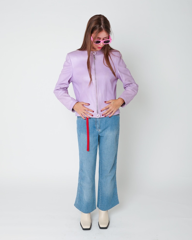 May include: A lavender leather jacket with a silver front zipper and a mandarin collar, paired with light blue denim jeans featuring a red fabric strip hanging from the waist and subtle embroidery at the hem, plus cream-colored ankle boots.