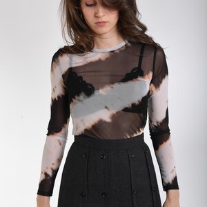 May include: A long-sleeve, sheer mesh top featuring a diagonal tie-dye pattern in black, white, and tan, worn over a black lace-trimmed bralette, paired with a dark gray, high-waisted, pleated skirt adorned with multiple black buttons.