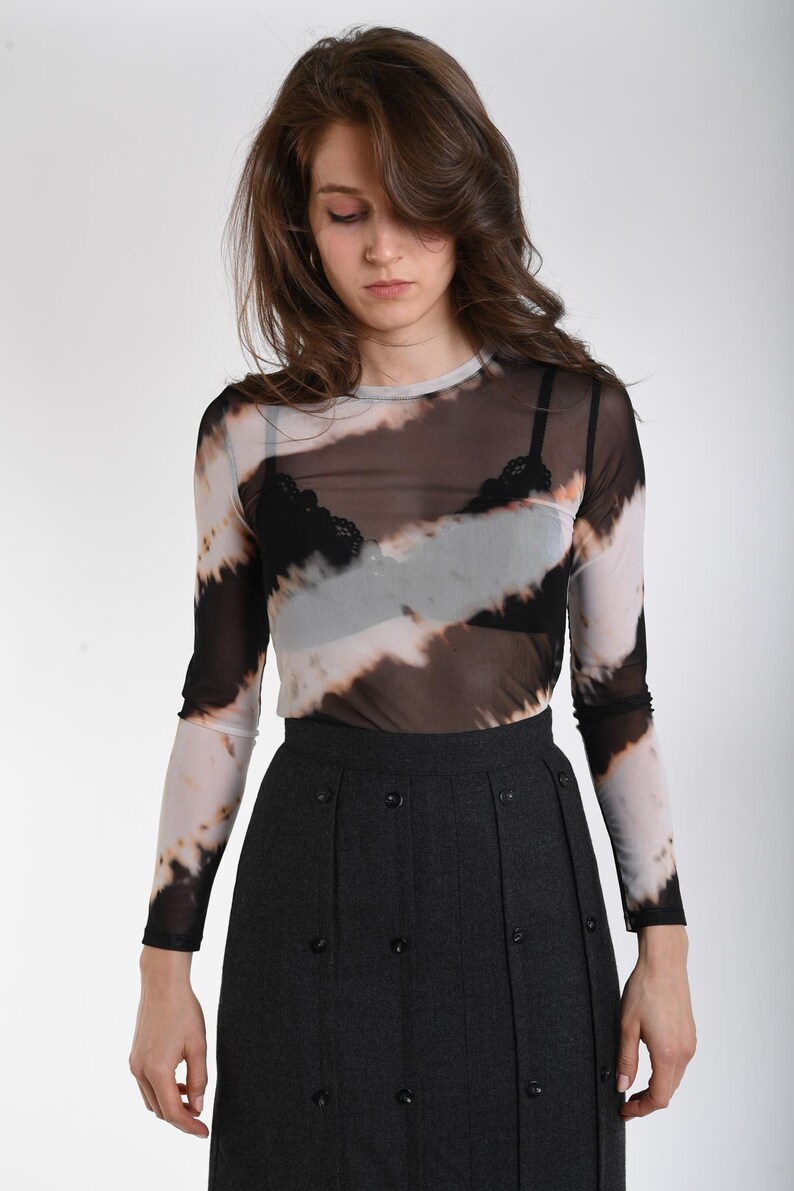 May include: A long-sleeve, sheer mesh top featuring a diagonal tie-dye pattern in black, white, and tan, worn over a black lace-trimmed bralette, paired with a dark gray, high-waisted, pleated skirt adorned with multiple black buttons.